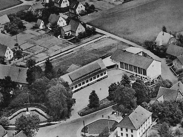 huellhorst-alte-schule-02.jpg Luftaufnahme eines historischen Schulgebäudes umgeben von typischen Wohnhäusern und viel Grünfläche.