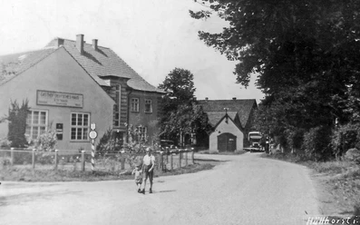 huellhorst-altes-spritzenhaus.jpg Historisches Gebäude mit Fachwerk und Satteldach, umgeben von Bäumen und Blumen, bei klarem Himmel.