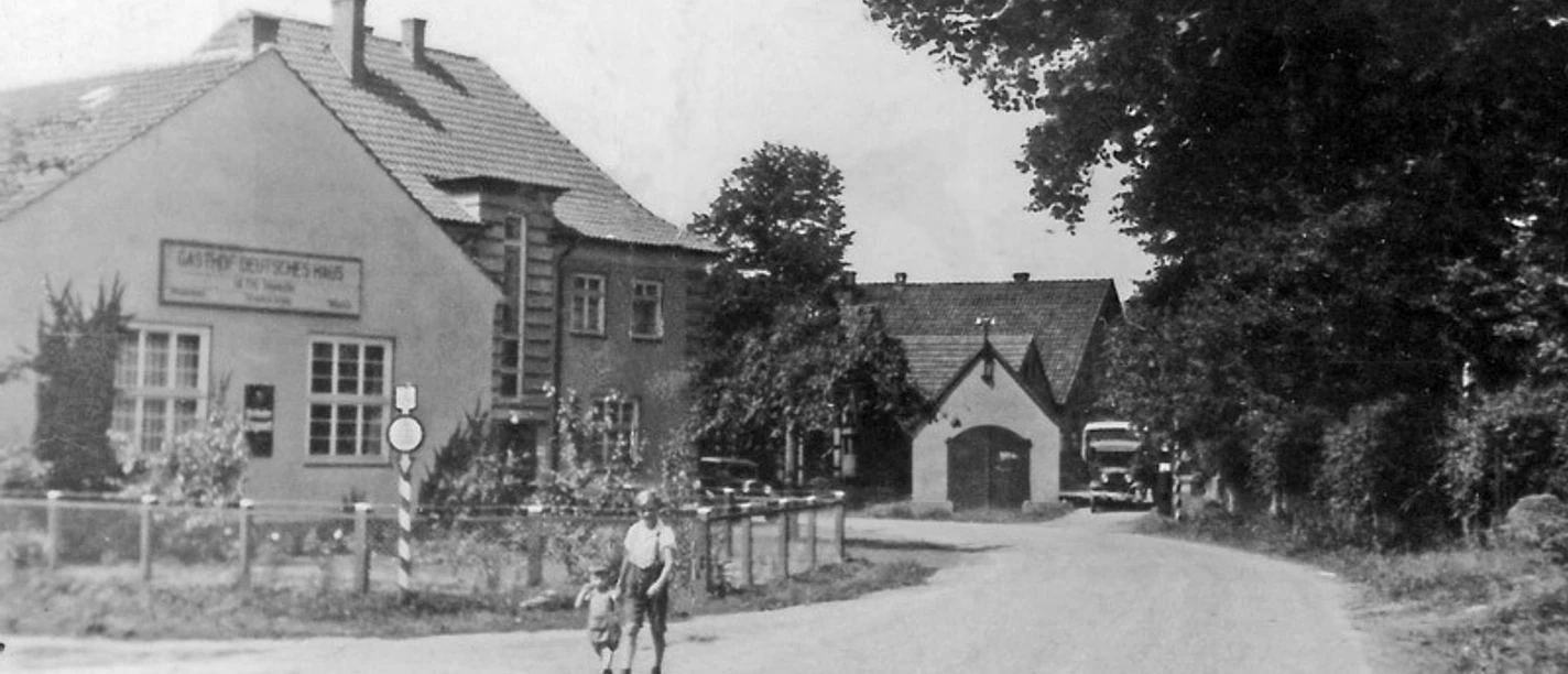 huellhorst-altes-spritzenhaus.jpg Historisches Gebäude mit Fachwerk und Satteldach, umgeben von Bäumen und Blumen, bei klarem Himmel.