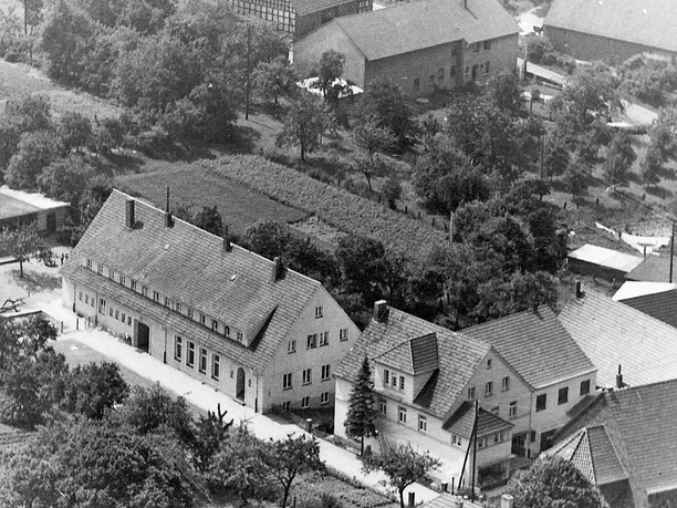 Luftaufnahme eines kleinen Dorfplatzes mit Fachwerkhäusern, grünen Bäumen und gepflegten Gärten.