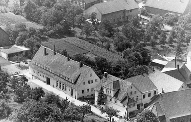 Dorfplatz.jpg Luftaufnahme eines kleinen Dorfplatzes mit Fachwerkhäusern, grünen Bäumen und gepflegten Gärten.