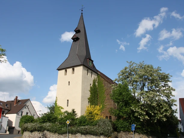 Die Andreaskirche Hüllhorst mit spitzem Kirchturm, umgeben von grünen Bäumen und blauem Himmel.