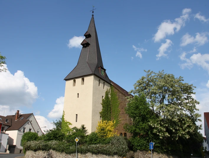 Die Andreaskirche Hüllhorst mit spitzem Kirchturm, umgeben von grünen Bäumen und blauem Himmel.