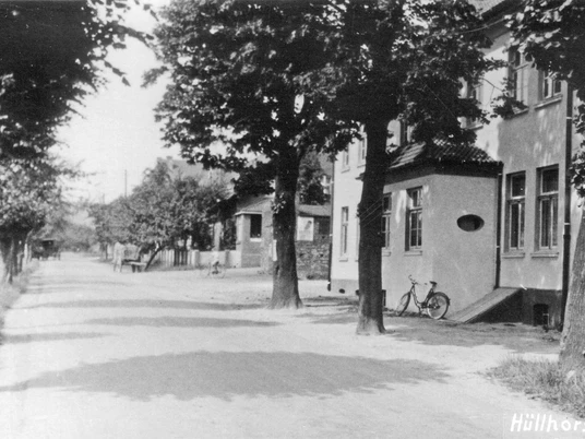 huellhorst-alte-hauptstraße.jpg Eine von Bäumen gesäumte Straße mit seitlich parkendem Fahrrad vor einem Backsteingebäude in Hüllhorst.