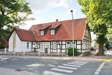 huellhorst-hauptstraße.jpg Fachwerkhaus mit roten Ziegeldach, umgeben von Bäumen, an einer ruhigen Straße mit Zebrastreifen.