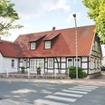 huellhorst-hauptstraße.jpg Fachwerkhaus mit roten Ziegeldach, umgeben von Bäumen, an einer ruhigen Straße mit Zebrastreifen.
