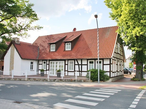 huellhorst-hauptstraße.jpg Fachwerkhaus mit roten Ziegeldach, umgeben von Bäumen, an einer ruhigen Straße mit Zebrastreifen.