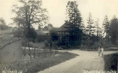 huellhorst-bad-lusmuehle.jpg Schwarz-weiß-Foto einer ländlichen Straße mit zwei gehenden Personen, hohen Bäumen und einem Gebäude.