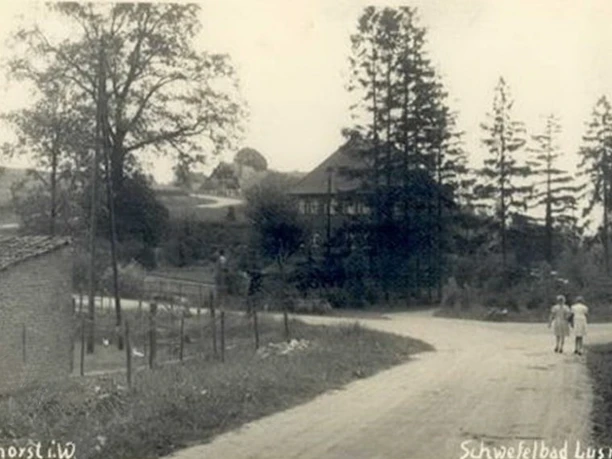 huellhorst-bad-lusmuehle.jpg Schwarz-weiß-Foto einer ländlichen Straße mit zwei gehenden Personen, hohen Bäumen und einem Gebäude.