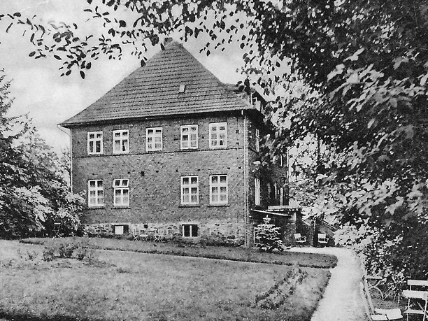 huellhorst-bad-lusmuehle-gasthof.jpg Historisches Backsteingebäude mit steilem Dach, umgeben von Bäumen und Wiese, im Hintergrund Wege.