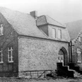 oberbauerschaft-dorfgemeinschaftshaus-alt.jpg Historisches Backsteingebäude mit Satteldach und Treppenstufen am Eingang, umgeben von kahlen Bäumen.