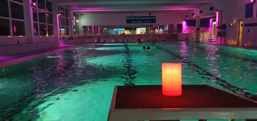 Ein Hallenbad mit stimmungsvoller Beleuchtung, mehrere Schwimmer im Wasser, und eine rote Kerze im Vordergrund.