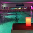 Ein Hallenbad mit stimmungsvoller Beleuchtung, mehrere Schwimmer im Wasser, und eine rote Kerze im Vordergrund.