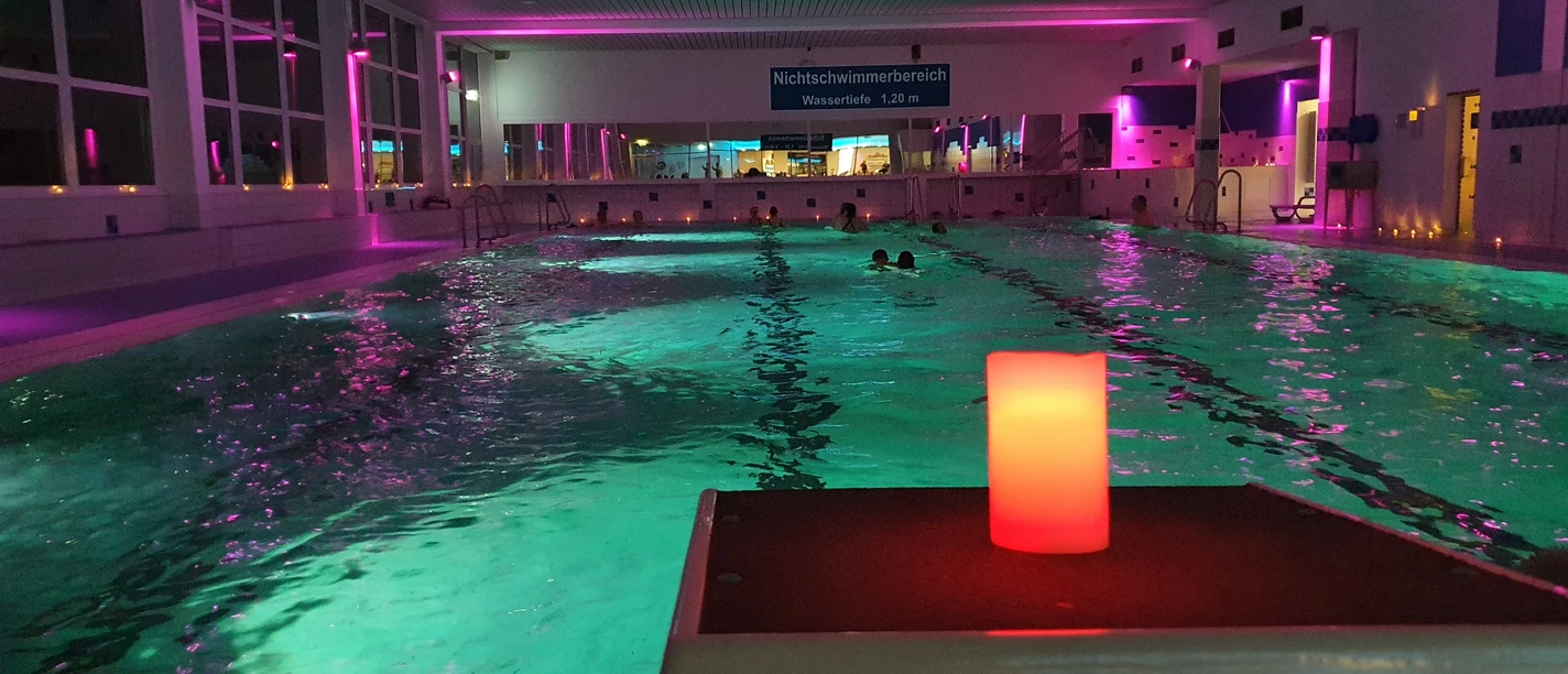 20230224_201951.jpg Ein Hallenbad mit stimmungsvoller Beleuchtung, mehrere Schwimmer im Wasser, und eine rote Kerze im Vordergrund.