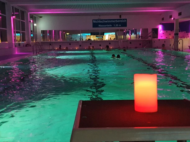 20230224_201951.jpg Ein Hallenbad mit stimmungsvoller Beleuchtung, mehrere Schwimmer im Wasser, und eine rote Kerze im Vordergrund.