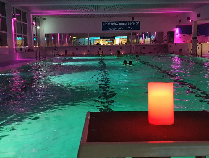 20230224_201951.jpg Ein Hallenbad mit stimmungsvoller Beleuchtung, mehrere Schwimmer im Wasser, und eine rote Kerze im Vordergrund.