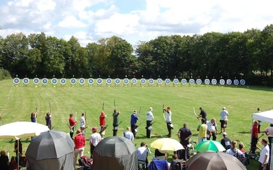 oberbauerschaft-alter-sportplatz-01.jpg Menschen beim Bogenschießen auf einer Wiese mit Zielscheiben, umgeben von Bäumen und Zuschauern.