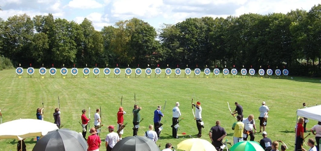 oberbauerschaft-alter-sportplatz-01.jpg Menschen beim Bogenschießen auf einer Wiese mit Zielscheiben, umgeben von Bäumen und Zuschauern.