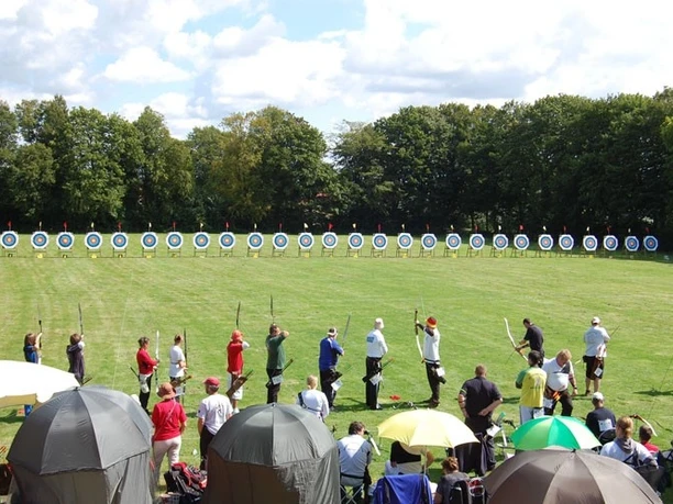 oberbauerschaft-alter-sportplatz-01.jpg Menschen beim Bogenschießen auf einer Wiese mit Zielscheiben, umgeben von Bäumen und Zuschauern.
