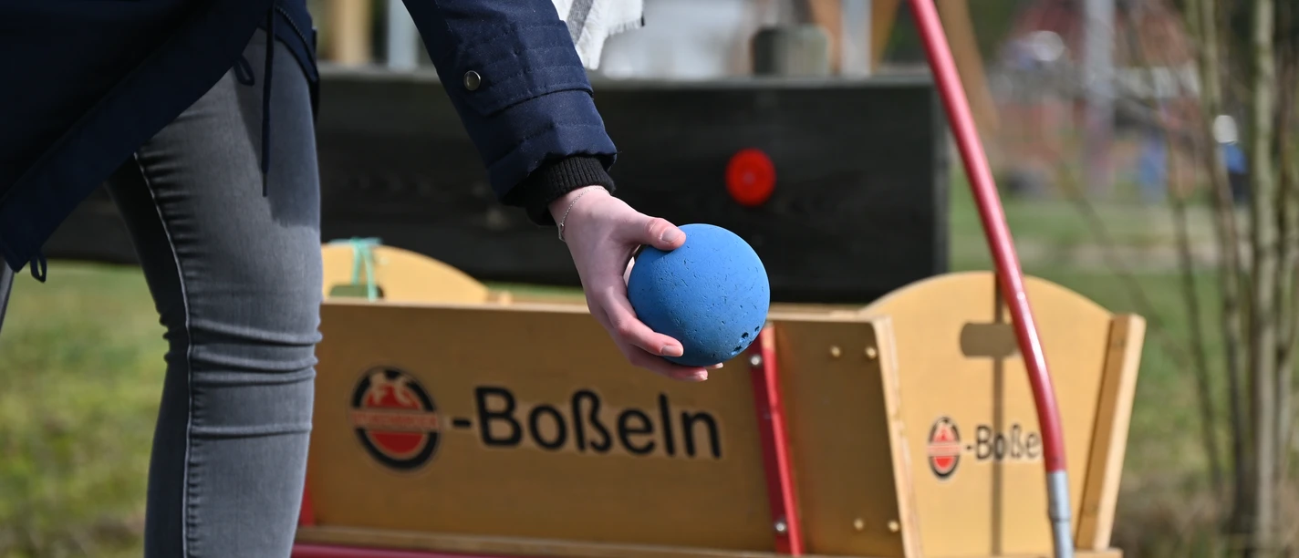 Boßeln in SHS_15_CC-BY-SA_Teutoburger_Wald_Stadt_Schloß_Holte-Stukenbrock.JPG Person hält blaue Boßelkugel vor einem Handwagen mit Boßeln-Schriftzug in Schloß Holte-Stukenbrock.