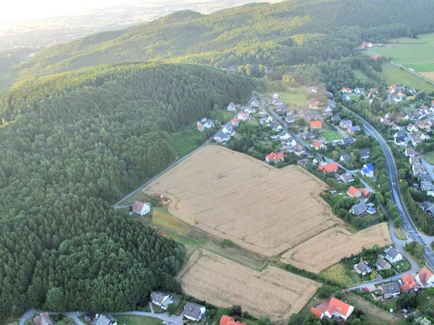 oberbauerschaft-koepperplatz-01.jpg Luftaufnahme eines Weilers mit Feldern und dichtem Wald, umgeben von sanft geschwungenen Hügeln.
