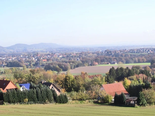 oberbauerschaft-augustus-hoffmann-bank-02.jpg Weitläufige Landschaft mit vereinzelten Häusern, umgeben von Feldern und Wäldern, im Hintergrund Hügel.