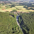 oberbauerschaft-wittekindsbruecke-01.jpg Luftaufnahme eines Dorfes, umgeben von dichten Wäldern und Feldern, mit Straßenverlauf im Vordergrund.