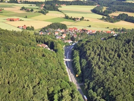 oberbauerschaft-wittekindsbruecke-01.jpg Luftaufnahme eines Dorfes, umgeben von dichten Wäldern und Feldern, mit Straßenverlauf im Vordergrund.