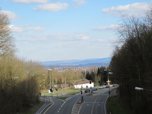 oberbauerschaft-wittekindsbruecke-02.JPG Straßenkreuzung vor bewaldeter Hügellandschaft, im Hintergrund eine weite Aussicht mit blauen Himmel.
