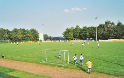 oberbauerschaft-sportplatz-festplatz-01.jpg Grüner Fußballplatz mit spielenden Teams, umgeben von Bäumen unter klarem Himmel und Flutlichtmasten.