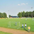 oberbauerschaft-sportplatz-festplatz-01.jpg Grüner Fußballplatz mit spielenden Teams, umgeben von Bäumen unter klarem Himmel und Flutlichtmasten.
