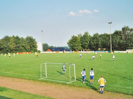 oberbauerschaft-sportplatz-festplatz-01.jpg Grüner Fußballplatz mit spielenden Teams, umgeben von Bäumen unter klarem Himmel und Flutlichtmasten.