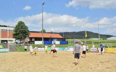 oberbauerschaft-sportplatz-festplatz-02.jpg Gruppe von Menschen spielt Beachvolleyball auf einem Sandplatz mit grüner Landschaft und Gebäuden im Hintergrund.