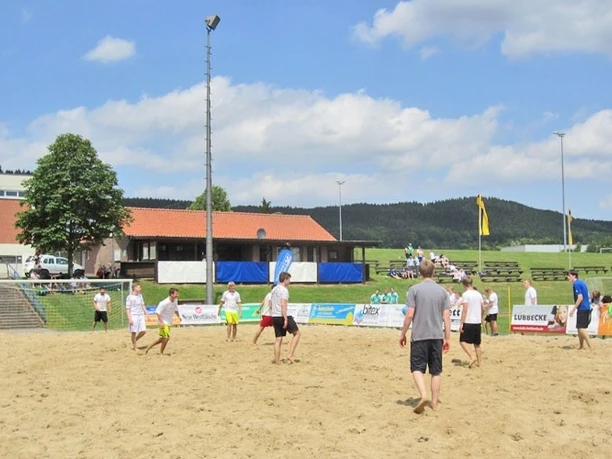 oberbauerschaft-sportplatz-festplatz-02.jpg Gruppe von Menschen spielt Beachvolleyball auf einem Sandplatz mit grüner Landschaft und Gebäuden im Hintergrund.