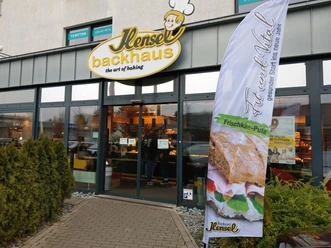 Eingang der Bäckerei Hensel mit großem Backhaus-Schild und Werbefahne vor der Glasfront