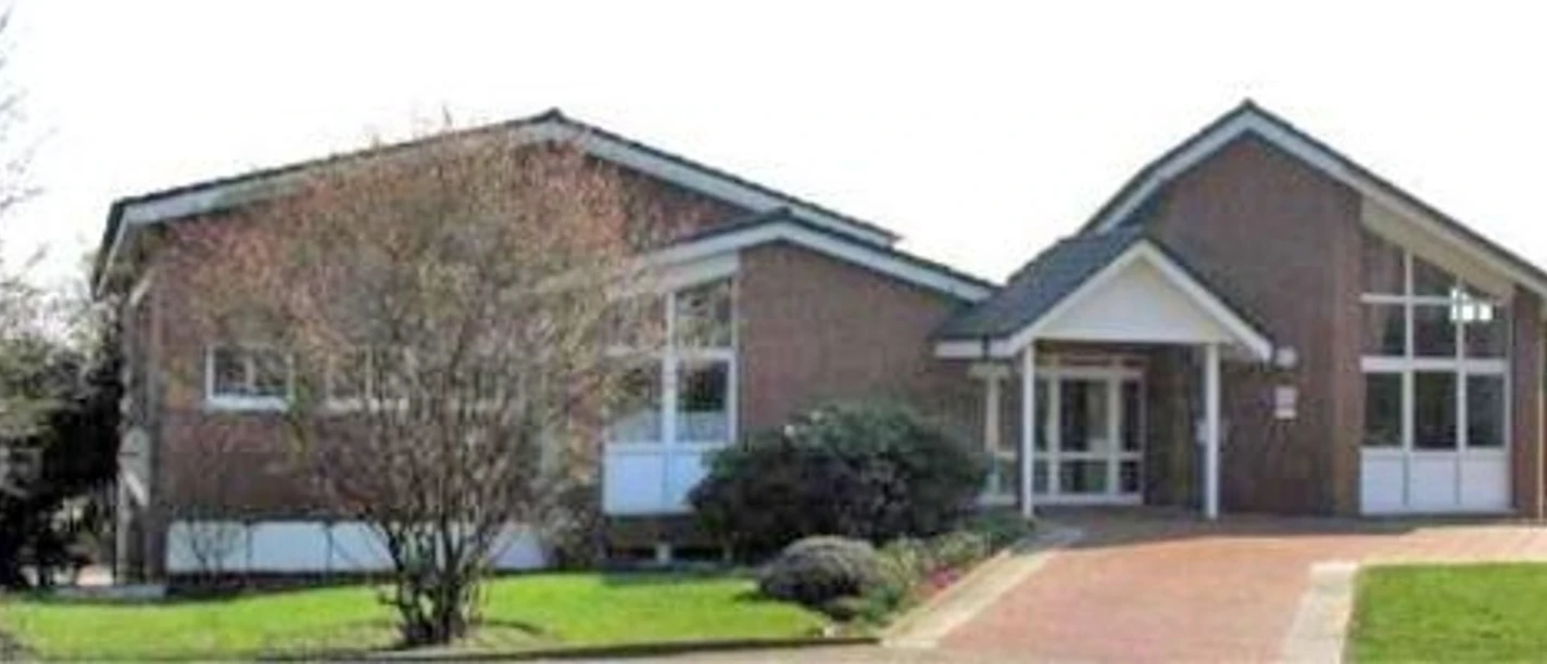 oberbauerschaft-gemeindehaus.jpg Backsteingebäude mit großen Fenstern, Eingang unter Vorbau, umgeben von Bäumen und gepflegtem Rasen.