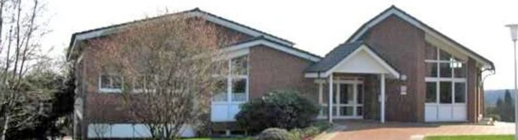 oberbauerschaft-gemeindehaus.jpg Backsteingebäude mit großen Fenstern, Eingang unter Vorbau, umgeben von Bäumen und gepflegtem Rasen.