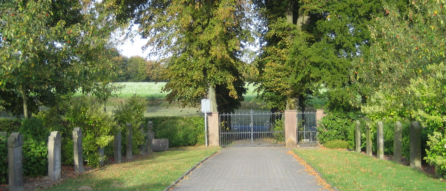 oberbauerschaft-friedhof-beendorf.JPG Eingang zu einem ländlichen Friedhof mit Tor und Bäumen, gesäumt von alten Grabsteinen.