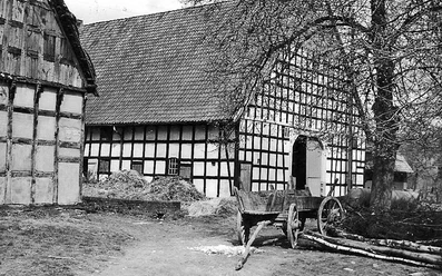 schnathorst-hof-wegener.jpg Schwarz-weißes historisches Fachwerkgebäude mit altem Holzkarren vor Strohballen und Bäumen.