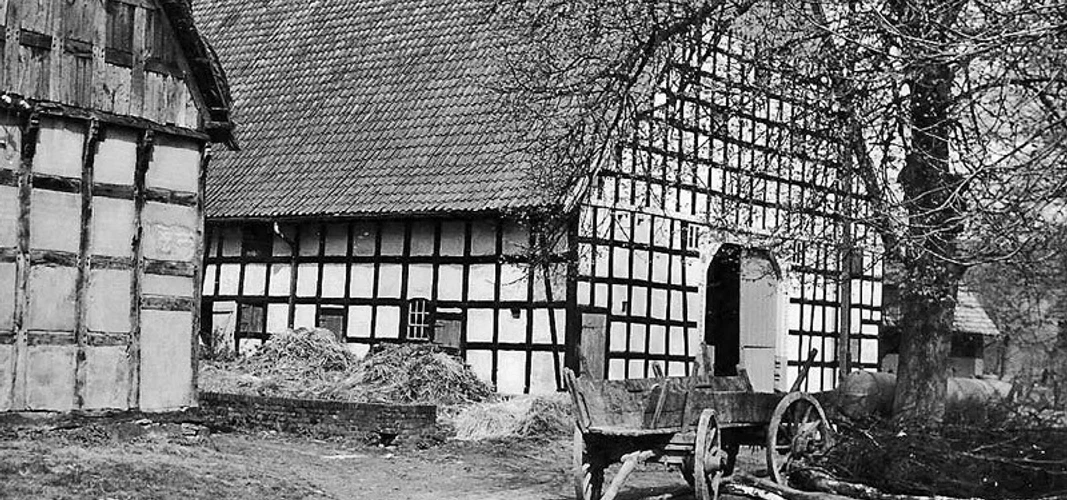 schnathorst-hof-wegener.jpg Schwarz-weißes historisches Fachwerkgebäude mit altem Holzkarren vor Strohballen und Bäumen.