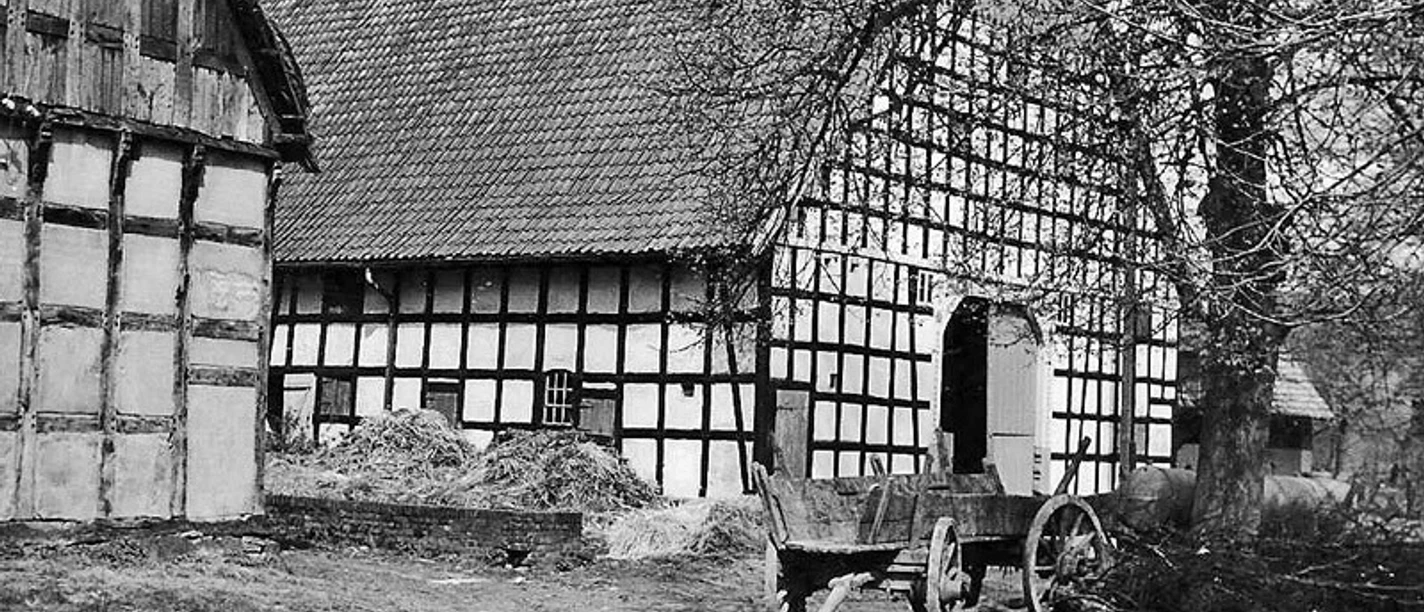 schnathorst-hof-wegener.jpg Schwarz-weißes historisches Fachwerkgebäude mit altem Holzkarren vor Strohballen und Bäumen.