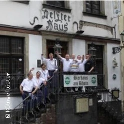 Personengruppe winkt fröhlich auf Treppe eines historischen Gasthauses mit Biergarten.