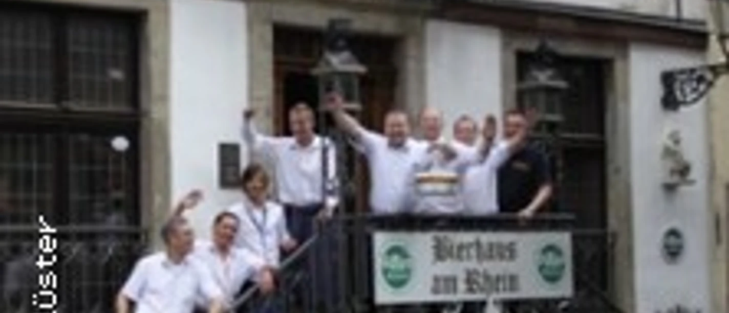 Personengruppe winkt fröhlich auf Treppe eines historischen Gasthauses mit Biergarten.