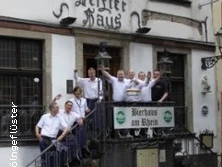 Personengruppe winkt fröhlich auf Treppe eines historischen Gasthauses mit Biergarten.