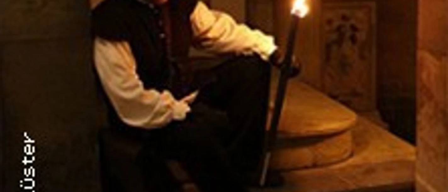 Ein im Mittelalter-Gewand gekleideter Mensch sitzt mit einer Fackel auf mittelalterlich anmutenden Stufen.