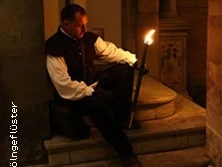 Ein im Mittelalter-Gewand gekleideter Mensch sitzt mit einer Fackel auf mittelalterlich anmutenden Stufen.