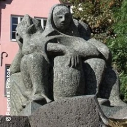 Steinernes Denkmal dreier sitzender, verschleierter Figuren mit ernstem Ausdruck auf einer Wiese.