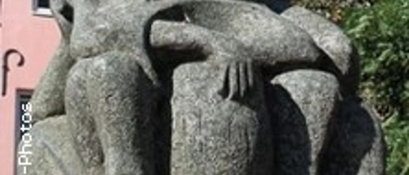 Steinernes Denkmal dreier sitzender, verschleierter Figuren mit ernstem Ausdruck auf einer Wiese.