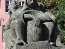 Steinernes Denkmal dreier sitzender, verschleierter Figuren mit ernstem Ausdruck auf einer Wiese.