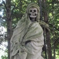 Verwitterte Skelettfigur aus Stein in Gewand, umgeben von dichtem Grün